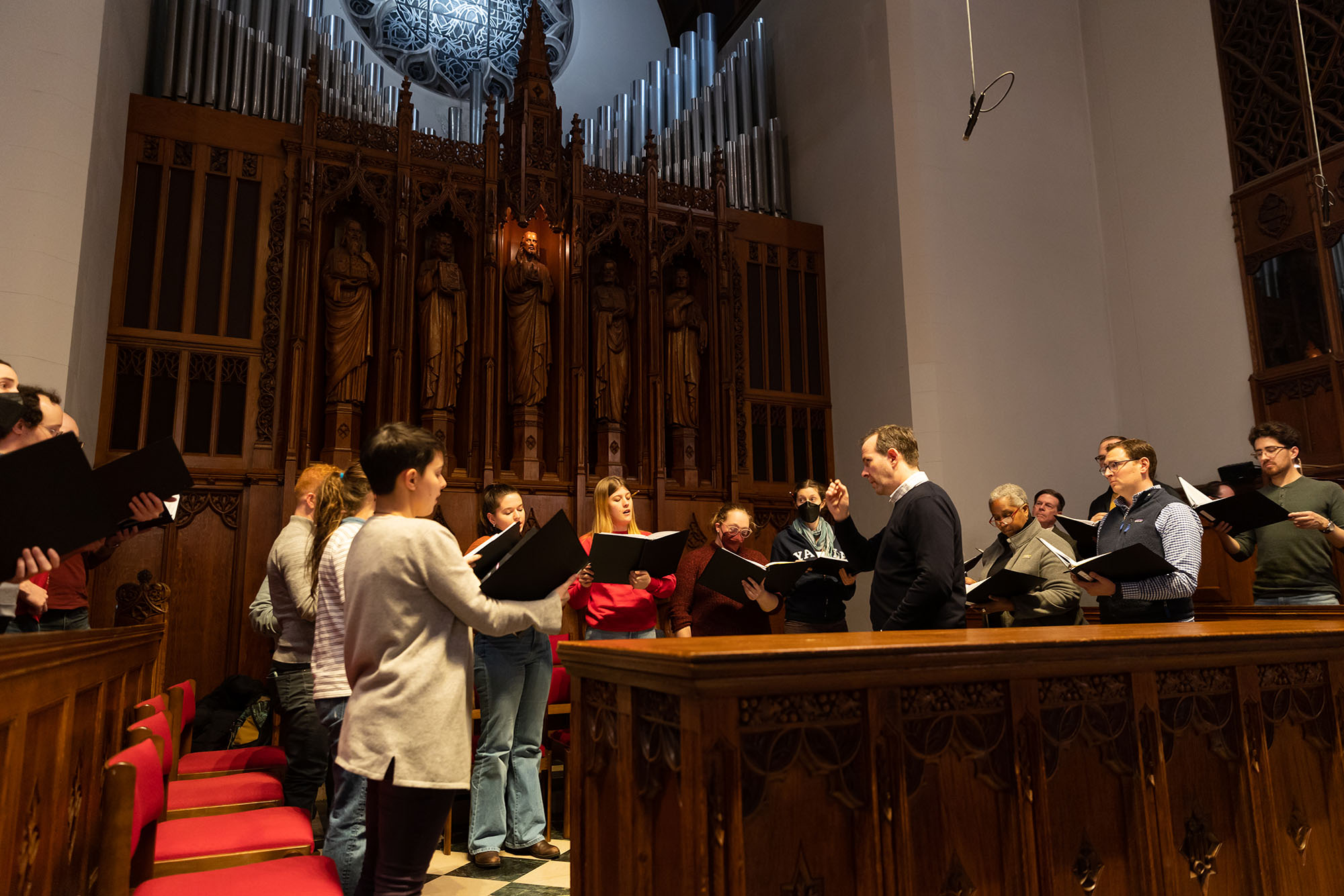 Photo: The Marsh Chapel choir practices on Monday, December 5, 2022. A group of people standing in a chapel hold music books and sing. A conductor stands in the middle of the half circle and leads.