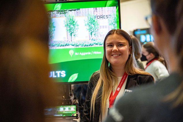 Photo: Emily Burbo (COM’24) presents her project on tree canopies at the Cross College Challenge exhibition in Questrom on Tuesday, Dec. 13. A young white woman with long blonde hair and wearing a white blouse a white blouse and black blazer, smiles as she stands in front of a large screen showing an example of a pop up forest.