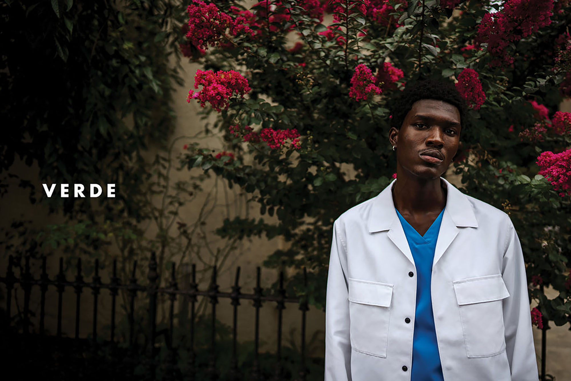 Photo: A young dark-skinned Black man models blue scrubs and a white lab coat in front of a large rose bush. The name "VERDE" is shown to the left of him in bold, white letters.
