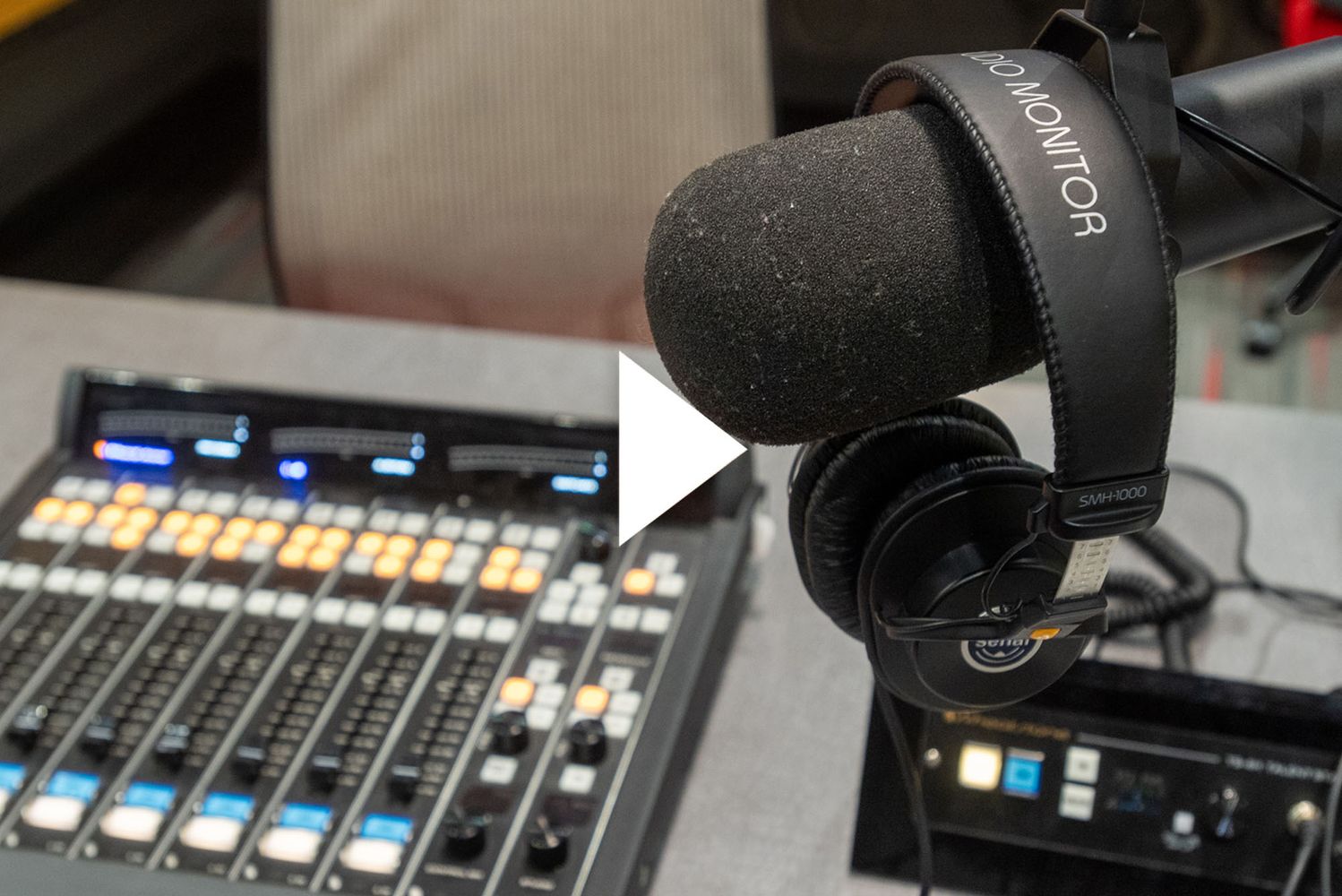 Photo: COM’s new renovated podcast studio. A grey table features a large black mixer, microphone, and professional grade headphones in a podcast studio recording room. A white triangle play button is overlaid in the center.