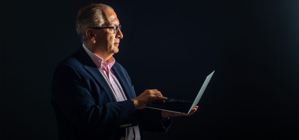 Azer Bestavros, wearing a suit against a black background, holds a laptop
