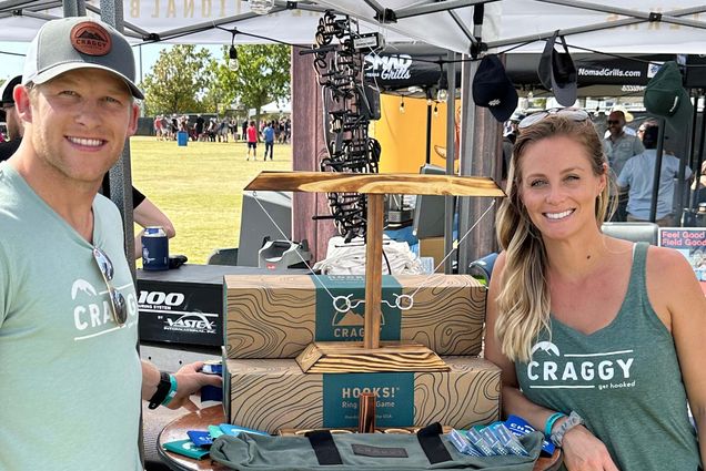 Kyle and Tabi Czarnecki show off HOOKS! The demand for their handcrafted game caught them off-guard when demand blew up last year. A man and woman stand on opposite sides of a table holding a game that consists of a piece of wood with strings and hooks attached to it.