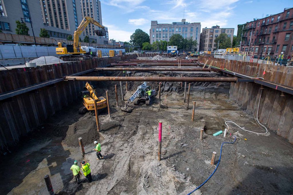 Photo: Construction site of the future Center for Computing and Data Sciences building. A large pit filled with metal work, pipes, and foundation work is shown being built in progress by construction workers.