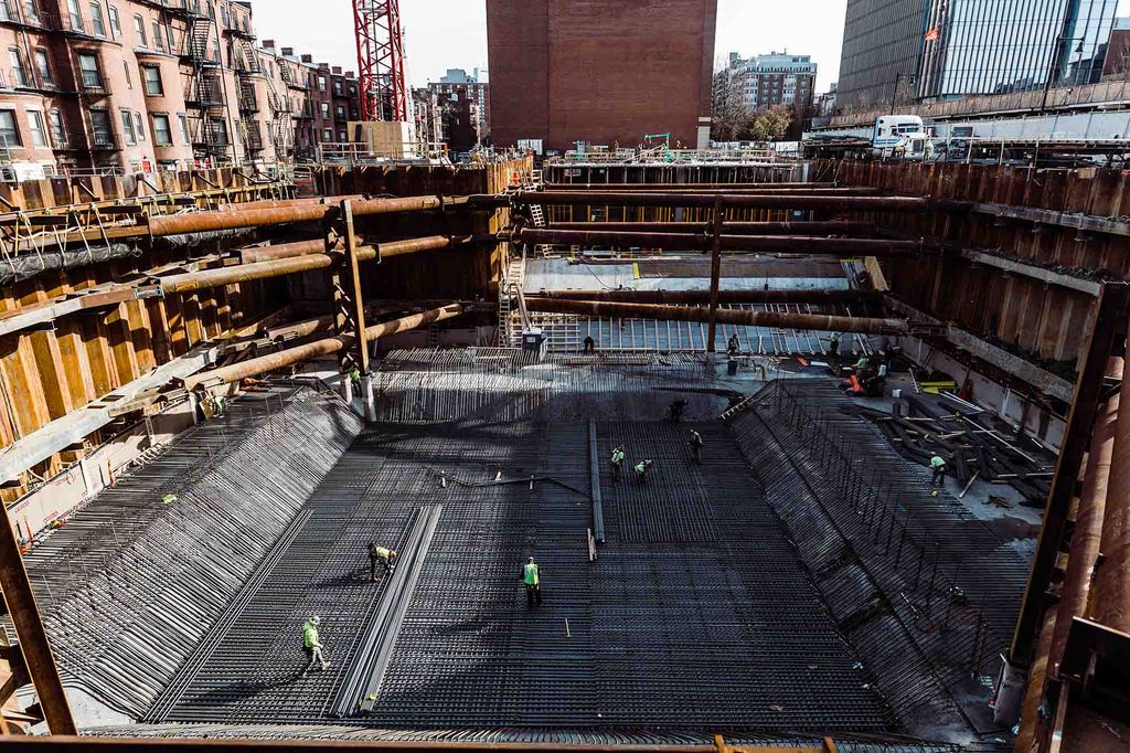 Photo: Construction site of the future Center for Computing and Data Sciences building. A large pit filled with metal work, pipes, and foundation work is shown being built in progress by construction workers.