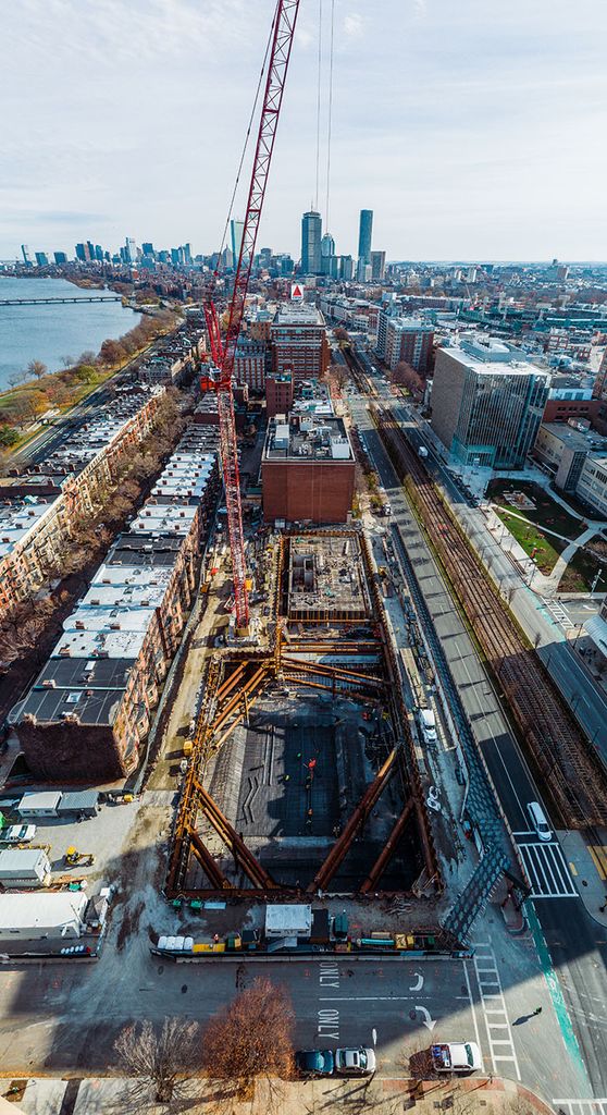 Photo: Construction site of the future Center for Computing and Data Sciences building. A large pit filled with metal work, pipes, and foundation work is shown being built in progress by construction workers.