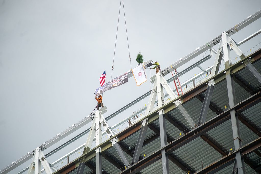 Photo: Covered in signatures, the final beam is put into place during the Topping Off ceremony at the Data Sciences Building September 30. The Marine flag and the American flag hang off the beam as two hard hat workers help lead it into place.