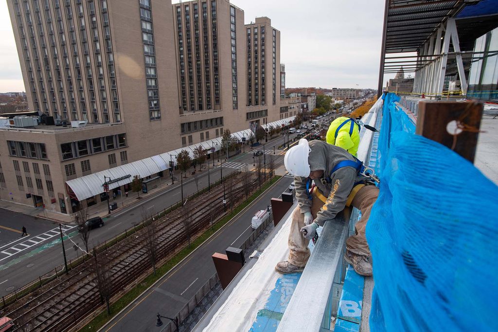 Photo of a construction worker sitting at the edge of a floor of the data sciences building and removing a blue tarp from the ground. Comm Ave is seen below him.