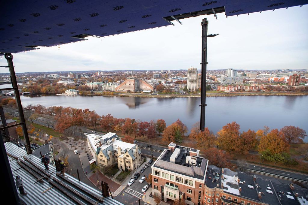 A large supporting beam is removed from the side of the data sciences building. Two construction workers look on from the platform below. In the background, the Cambridge side of the charles river is seen, and the trees along the river have fall-colored leaves.