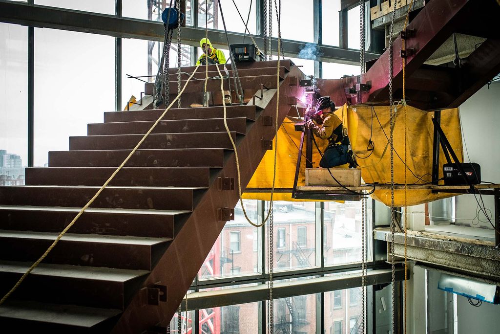 Photo: Installation begins on the staircase inside of The BU Center for Computing & Data Sciences on March 17, 2022. A construction worker welds in the dark on a staircase. They hang upwards on a small platform as they work on a high edge.