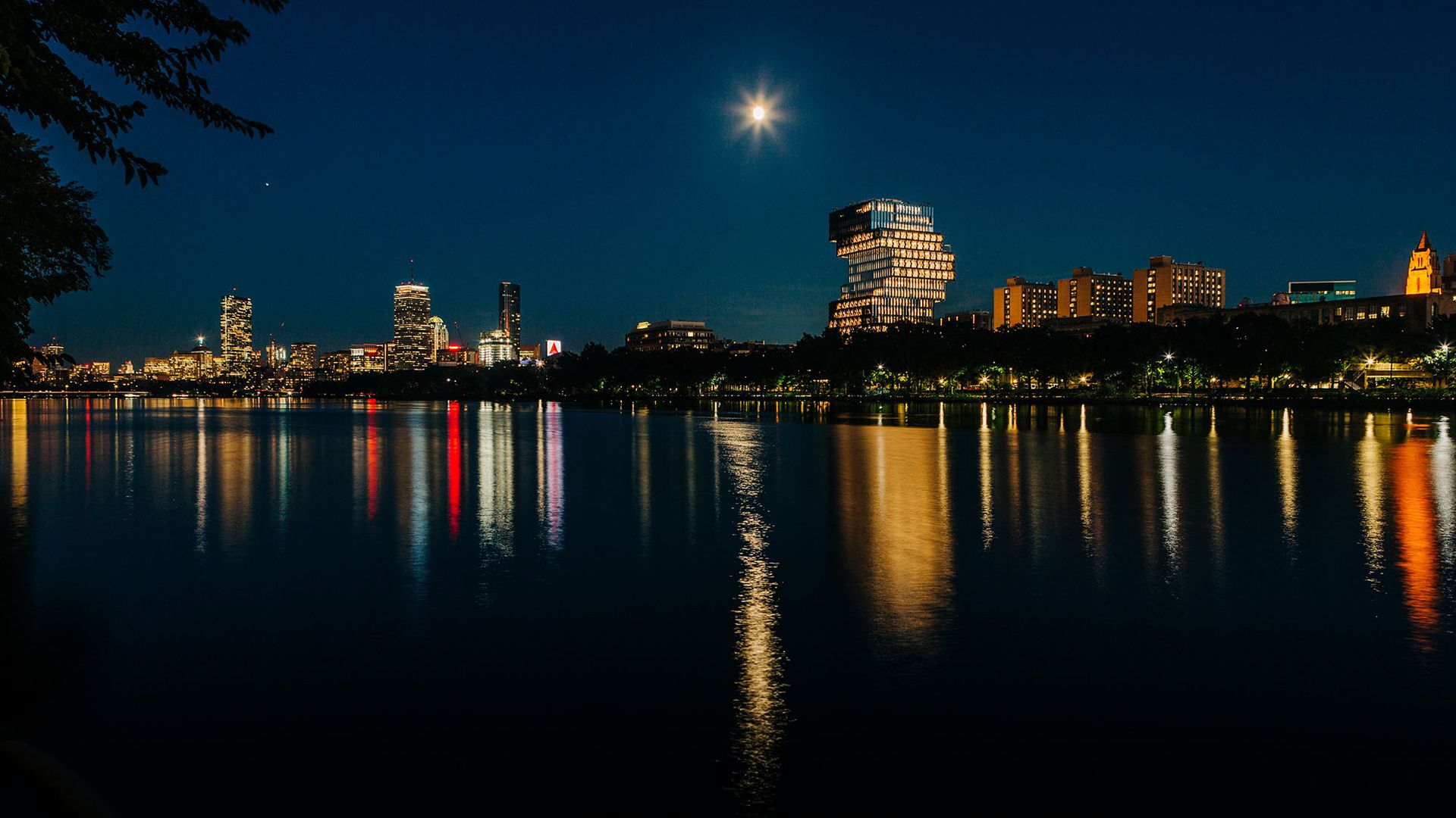 Photo: Boston University’s Center for Computing & Data Sciences is shown as part of the nighttime Boston Skyline. Buildings are lit and a reflection of the city is shown on the river in front of everything.