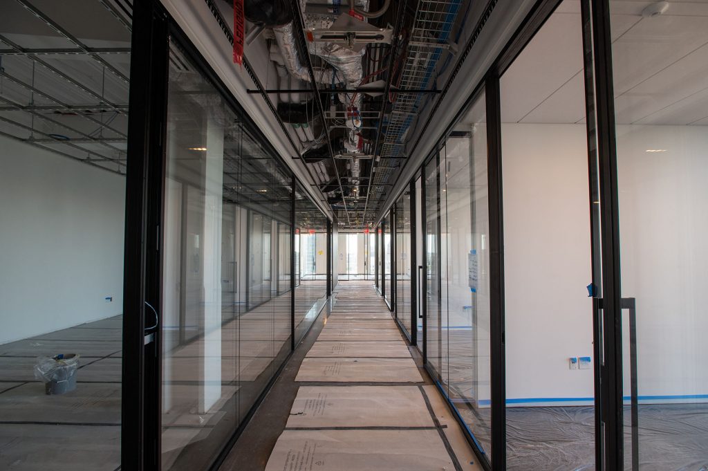 Photo: Interior of the Center for Computing and Data Science as work continues to be done. View of a hallway made of room rooms with sides and doors made of glass. A not finished floor is lined with cardboard.