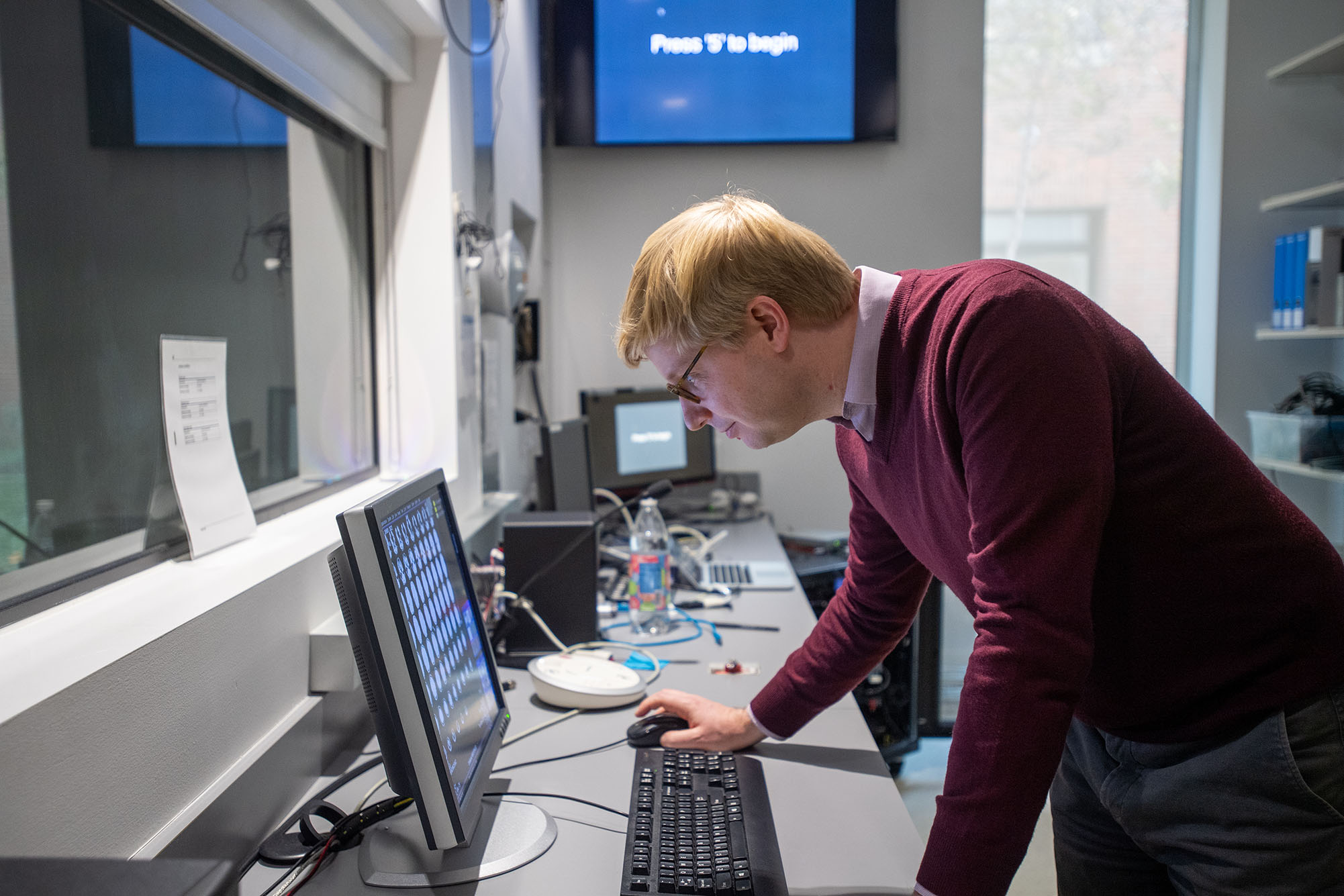 Photo: Tyler Perrachione, a Sargent associate professor, supervising the images from a student-designed experiment in the Cognitive Neuroimaging Center in CILSE. A white man wearing a burgundy sweater leans over and looks into a computer screen in a lab environment.