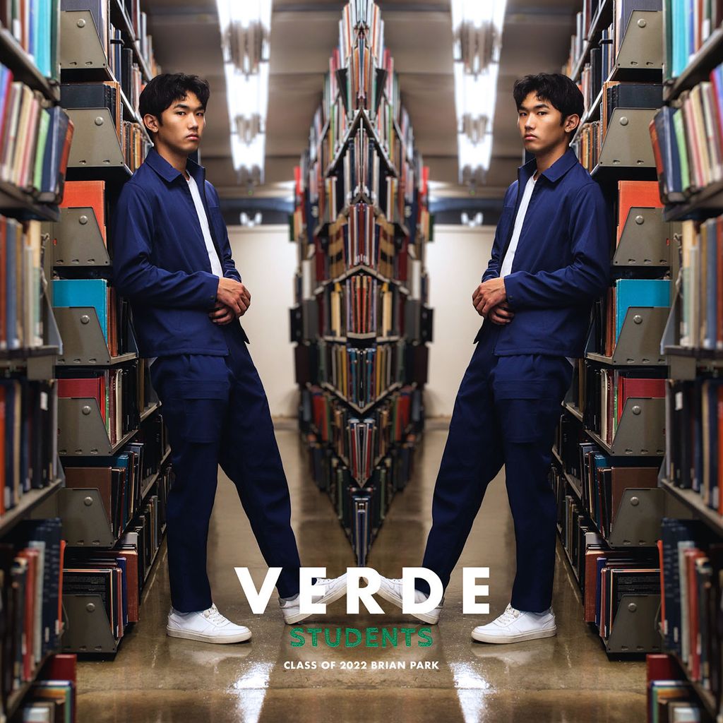 Image: Photo of Brian Park (Questrom’23) modeling VERDE scrubs for the #VERDEstudents campaign. A young Asian man wearing a white shirt, navy blue collared shirt and pants, and white sneakers poses in an aisle of a library. Photo is mirrored so the model is shown on both sides.