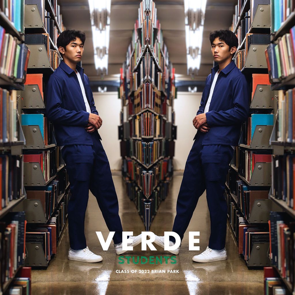 Image: Photo of Brian Park (Questrom’23) modeling VERDE scrubs for the #VERDEstudents campaign. A young Asian man wearing a white shirt, navy blue collared shirt and pants, and white sneakers poses in an aisle of a library. Photo is mirrored so the model is shown on both sides.