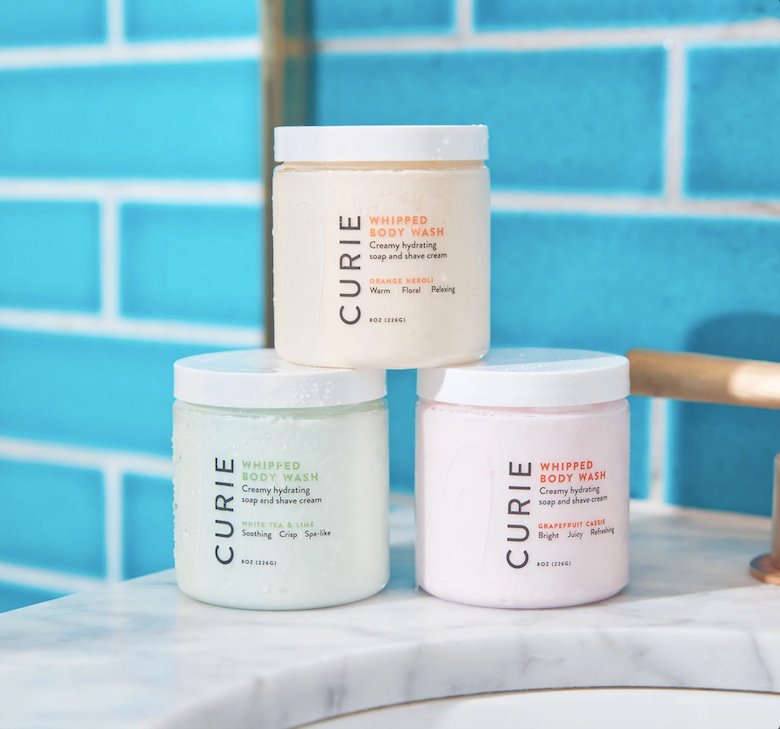 Photo: Display of three bath scrubs on a marble bathroom countertop. They are arranged in a mini pyramid with branding for the brand Curie.