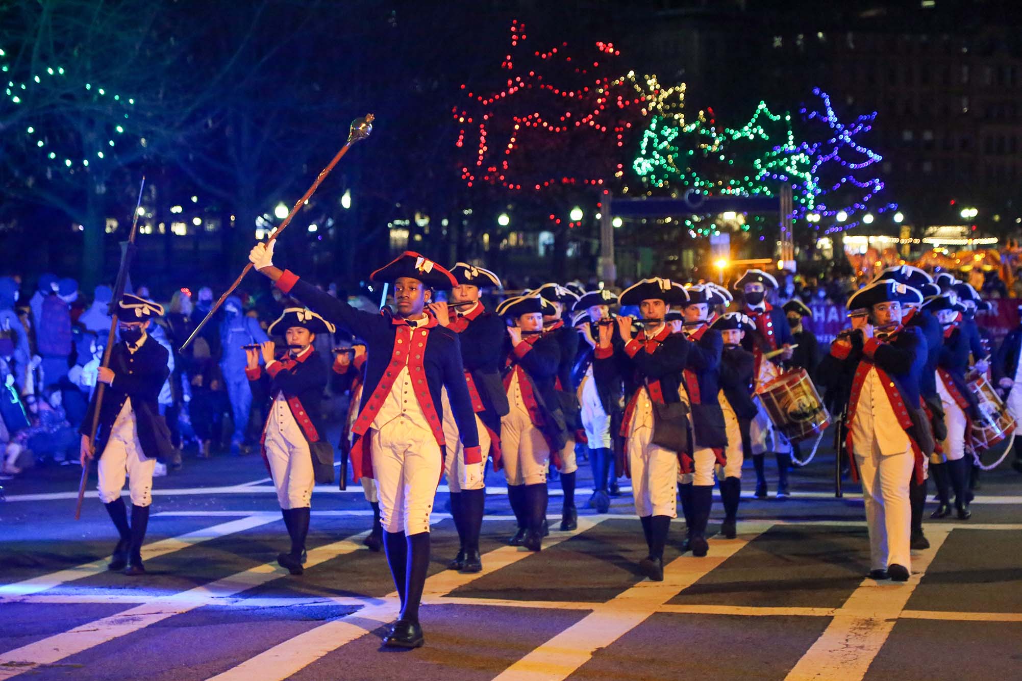 Photo: A group of young boys wearing old colonial uniforms march down a street lined with trees decorated with red, green, and yellow Christmas lights. The lead person holds up a long cane/baton while the other march in neat lines as they play various instruments.