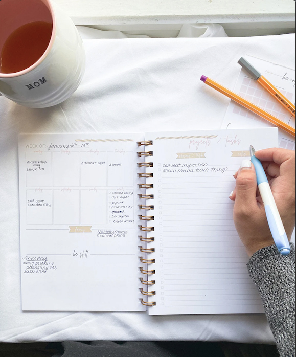Photo: Overhead shot of a planner laid open on a marble tabletop. An empty mug is placed to the left of the planner and a hand is shown writing in the planner.
