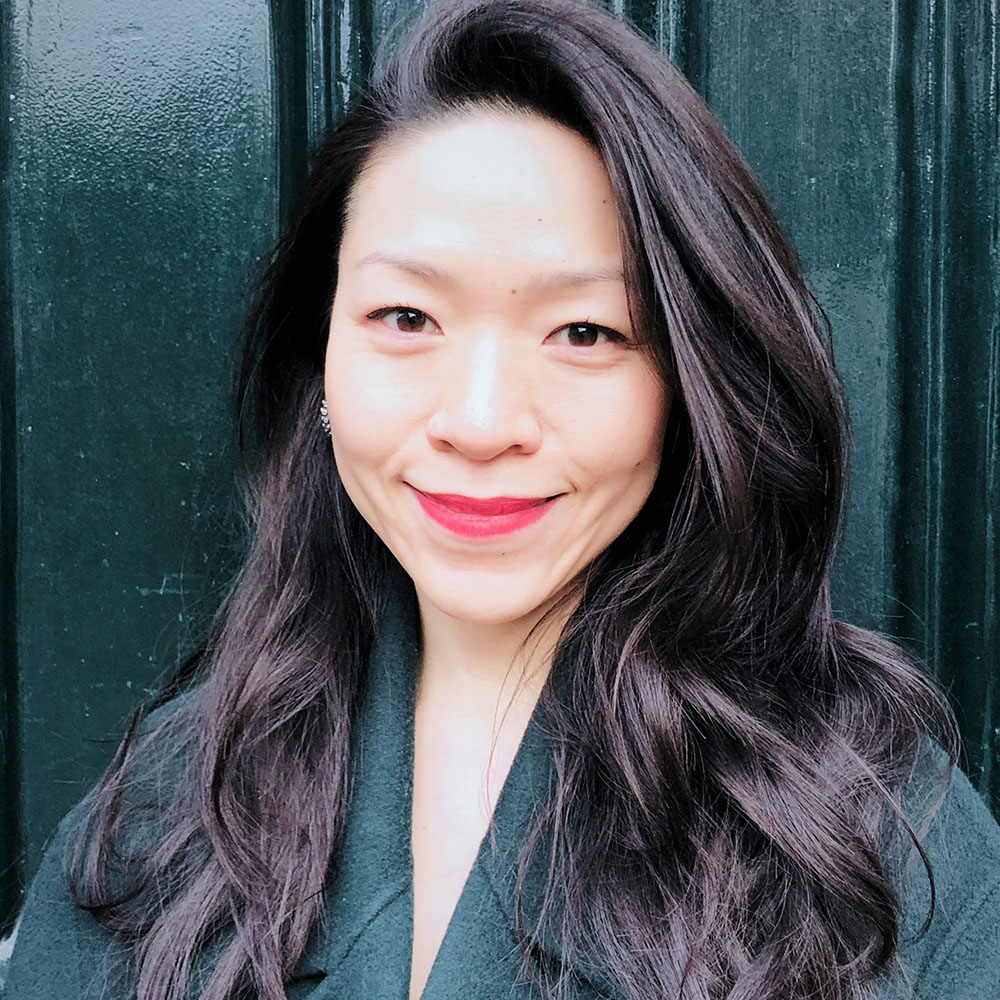 Photo: Headshot of Lewina O. Lee. An Asian woman with long, black hair and wearing coral red lipstick and green wool coat smiles for the camera.