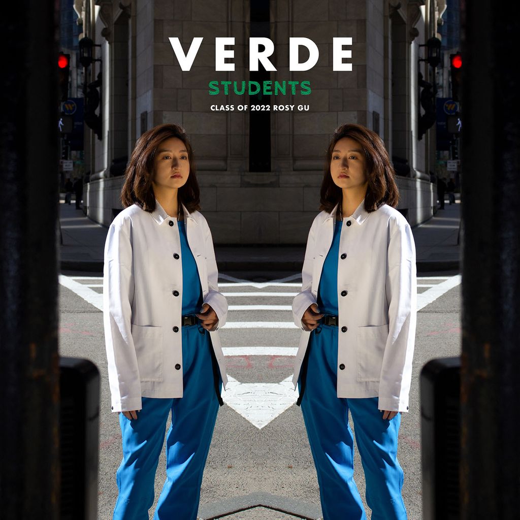 Image: Photo of Rosy Gu (COM’23) modeling VERDE scrubs for the #VERDEstudents campaign. A young Asian woman wearing blue scrubs and a white lab coat stands at a street crossing on a sunny day. The photo is mirrored to show the woman on both sides facing each other.