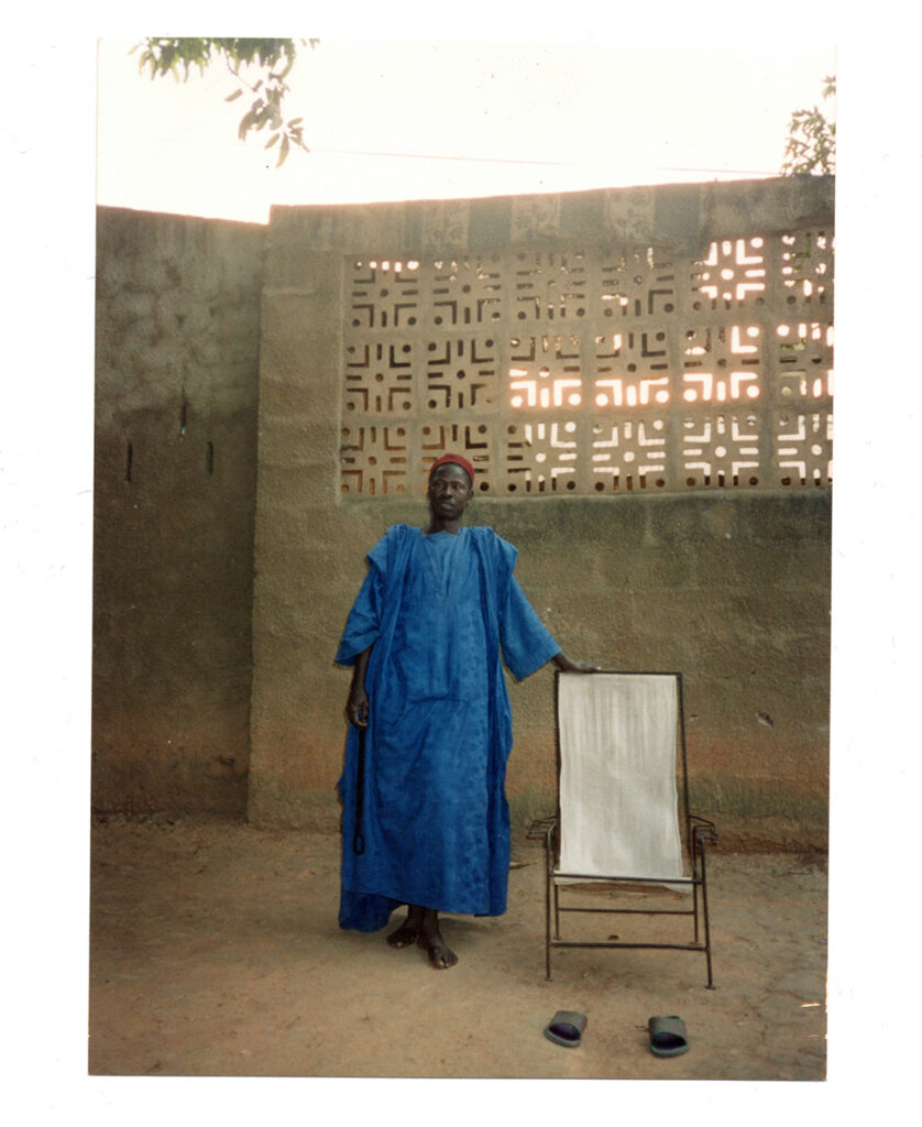 Photo: A Black man wearing a long blue African top, pants, and African print cap, leans against a small board with a large sheet of paper on it and stands in an empty area with a stone wall behind him.