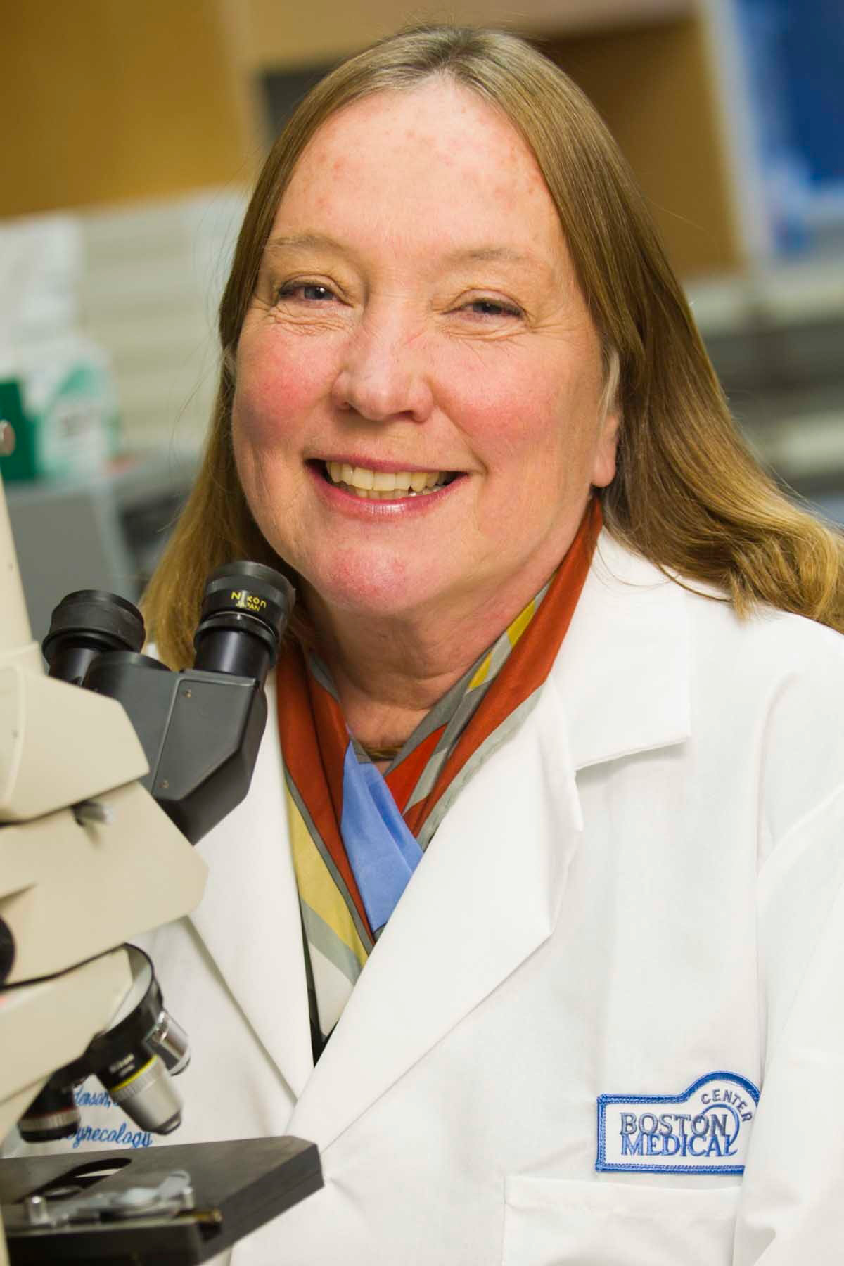 Photo: Deborah Anderson poses in a lab next to a microscope. A white woman with dirty blonde hair and wearing a lab coat smiles and poses next to a lab microscope.