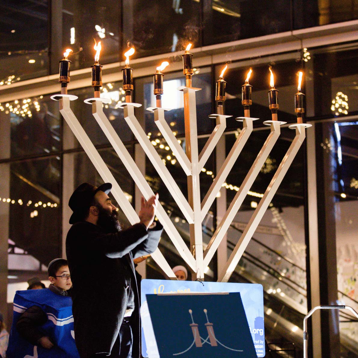 Two pictures. The one on the left features a bearded man with a brimmed hat on stands in front of a giant, fully lit Menorah. The one on the right features an indiviual capturing a lit Christmas tree with an iPhone. The individual is featured in the bottom left hand corner while the bottom portion of the Christmas tree is featured in the right hand corners.