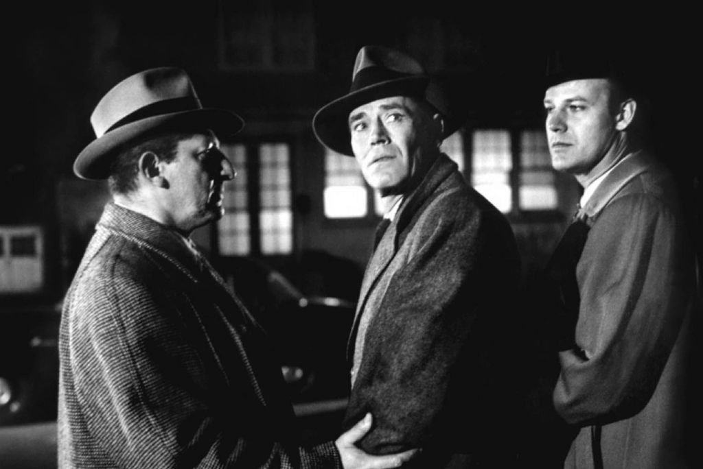 Alfred Hitchcock made his film as factual as he could, but still used his brilliant technique to make audiences feel the plight of The Wrong Man, played by Henry Fonda. Here Fonda stands between two white middle aged men who are also detectives. All men are wearing hats. Credit: cineclassico/Alamy Stock Photo