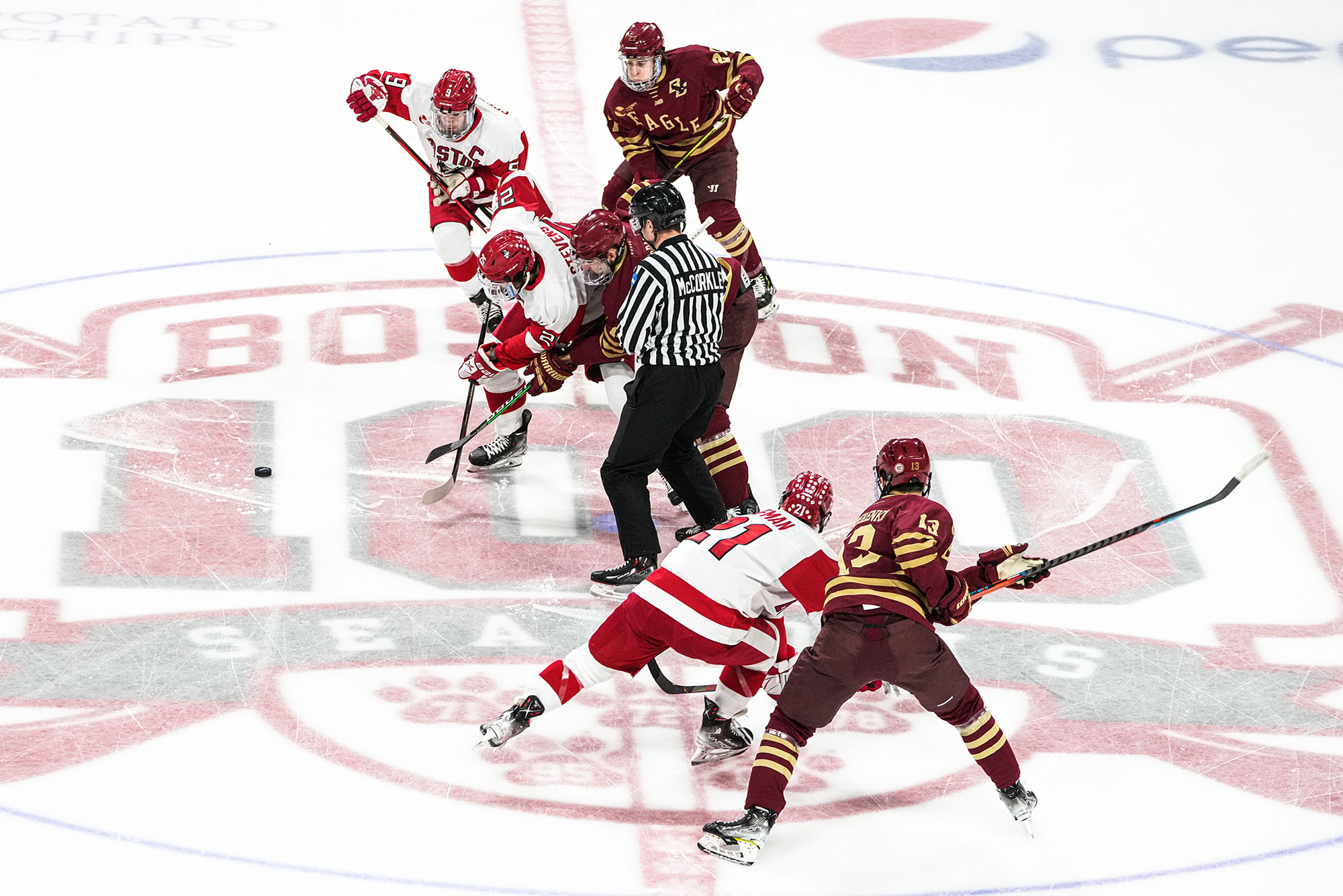 Photo: BU Men's ice hockey faces-off against BC. Various BU hockey players grapple and push on the ice against BC players to get the puck. A referee stands in the center of the group.