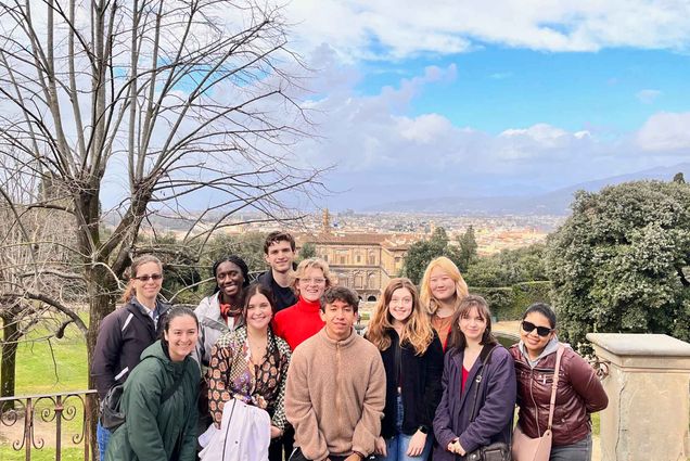 Photo: A group of students dressed in jackets and cold gear smile for the camera on a sunny, partly cloudy day. Distant buildings can be seen behind them.