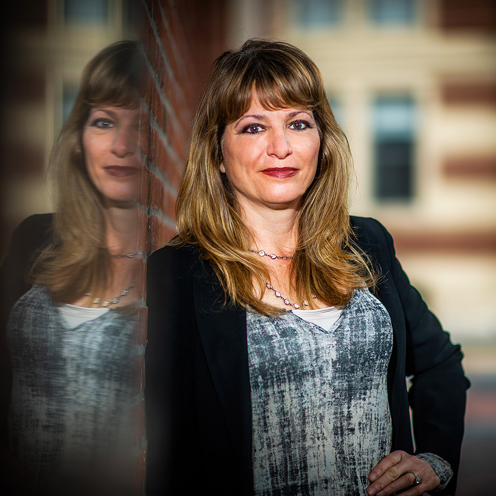 Photo: Belinda Borrelli poses for a photo. A white woman with dirty blonde hair and wearing red lipstick, a blue plaid blouse, and black cardigan, smiles and leans next to a reflective wall.