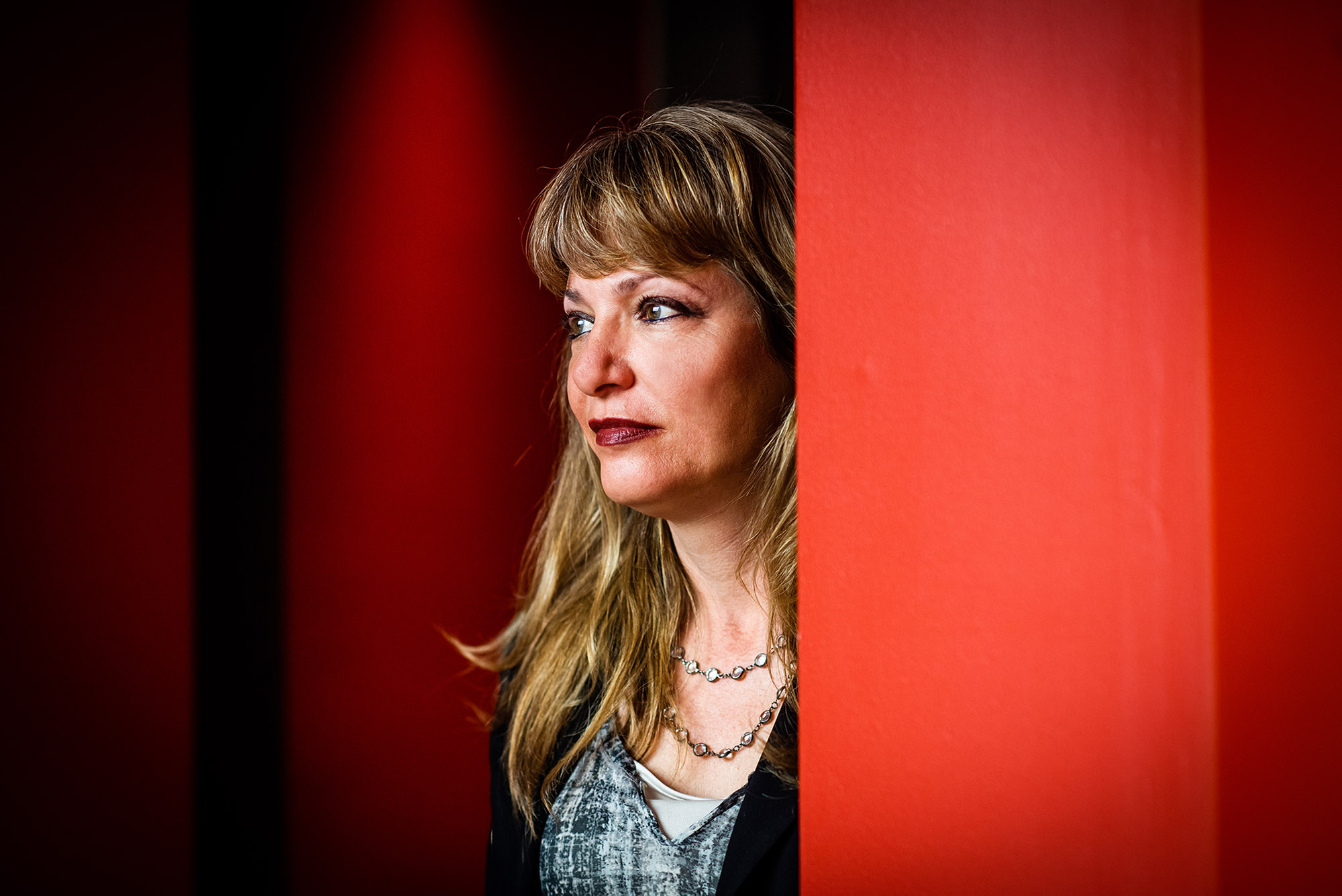 Photo: Belinda Borrelli poses for a photo. A white woman with dirty blonde hair and wearing red lipstick, a blue plaid blouse, and black cardigan, peers to the left as she stands partially behind a red wall.