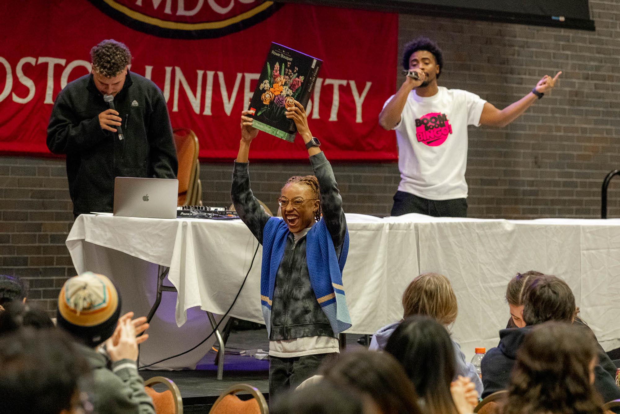Photo: Raquel Joseph, a young Black woman wearing glasses and a grey-themed tye-dye jumpsuit, laughs and holds up a large Lego flower set in glee. A DJ wearing a shirt that reads "Boom boom bingo!" speaks into a microphone at a white table behind her.