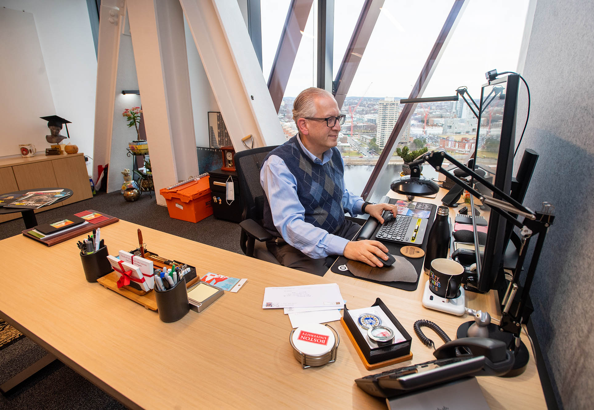 Photo: Professor and associate provost for computing and data science, Azer Bestavros, gets some work done in his new office in the Center for Computing & Data Sciences. An older man sits at a large wooden desk and works on his desktop. A large window wall to the right of him lights up the room.
