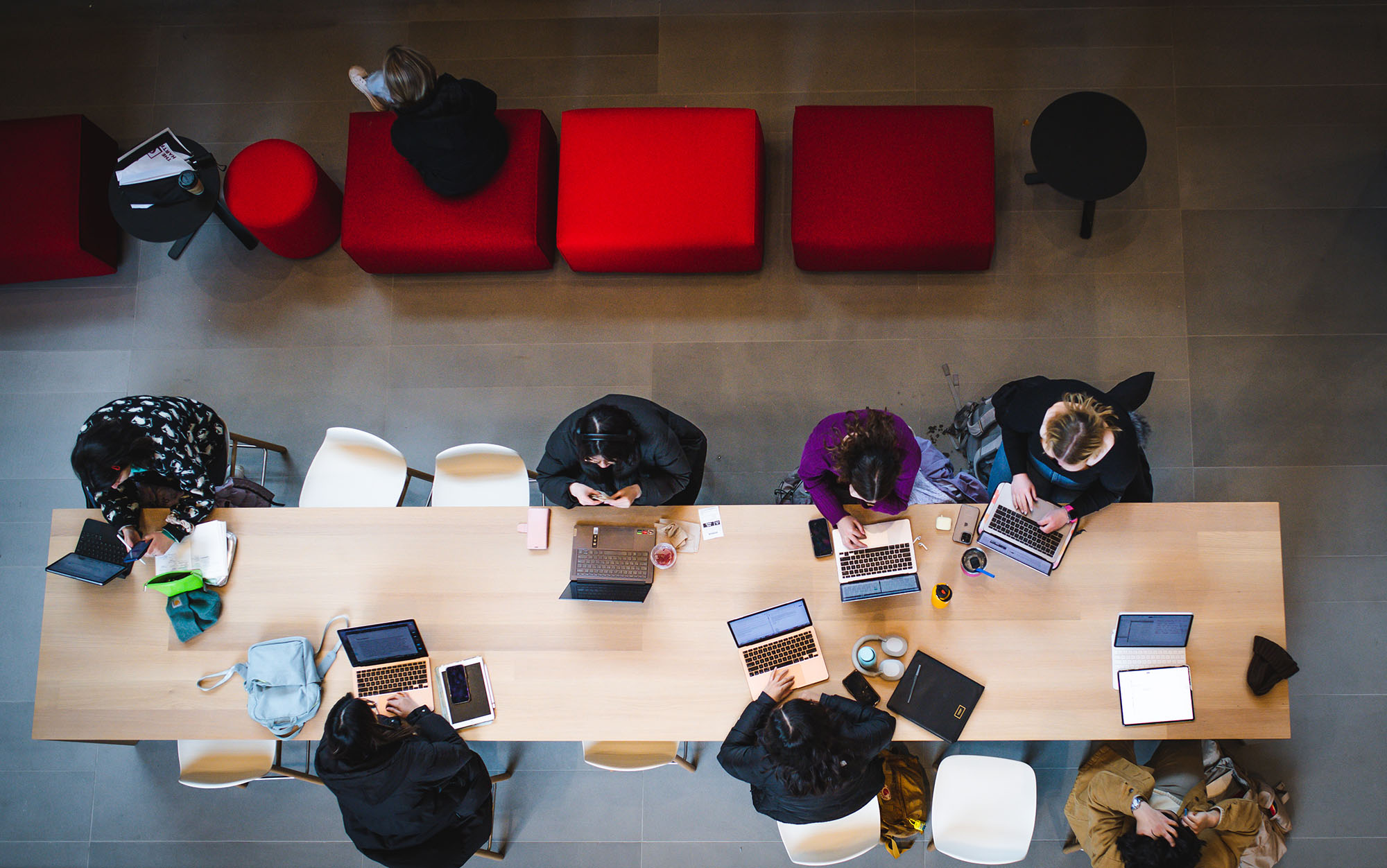 Photo: Over head shot of students gathered in the open spaces of the Center for Computing and Data Sciences. Various students sit and work on their laptops at a long wooden table.