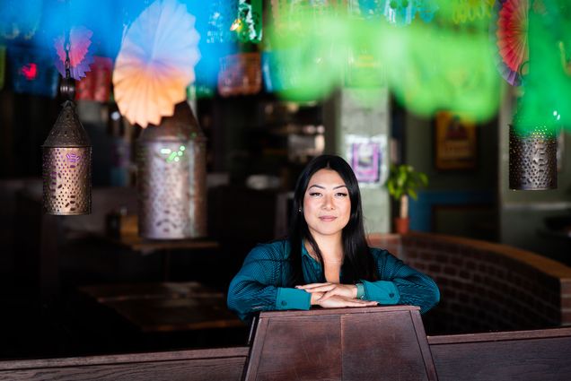 Photo: Businessperson Catarina Chang (SHA'11) poses for a photo at Sunset Cantina on January 17,2023. A young Asian woman wearing a long-sleeved jewell blue blouse, poses with arms folded over each other on a wooden divider. Wooden dining tables can be seen in a seating area behind her. The ceiling/walls are decorated with brightly colored fan ornaments of various sizes.