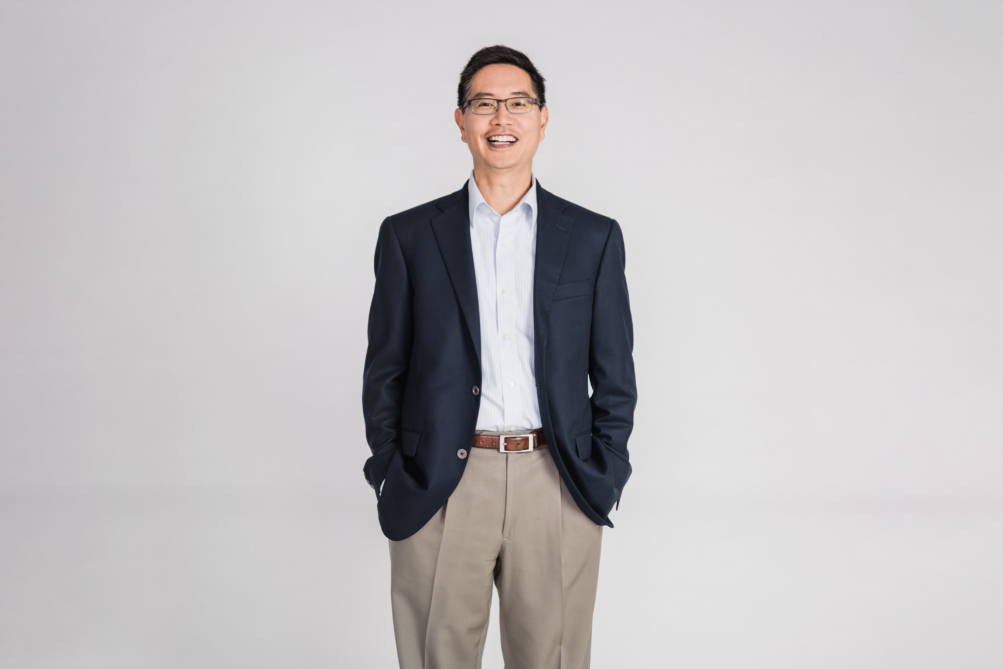 Photo of Chris Chen, an Asian American man with glasses who wears a blue suit jacket and smiles with his hands in his pockets. He stands in front of a light grey background.