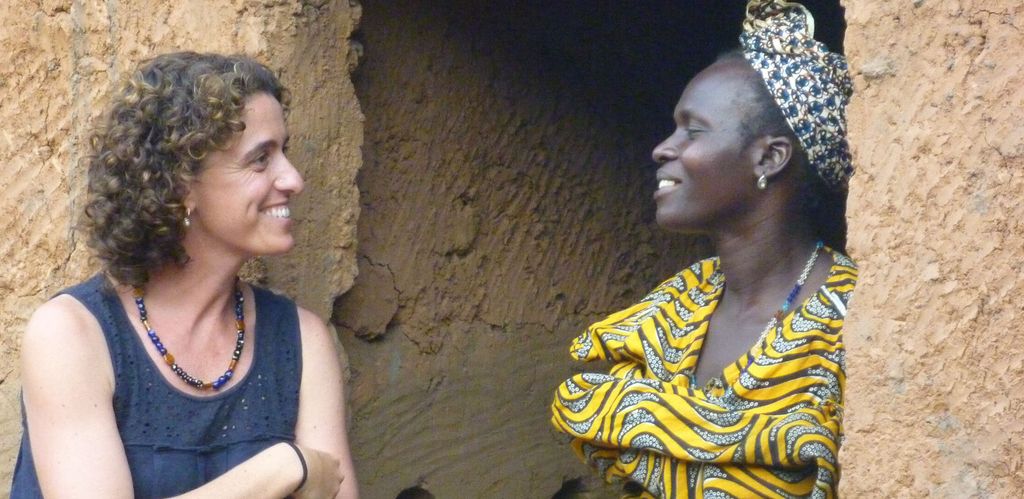 A white woman stands with arms crossed and smiles as she looks to her right to a Black woman wearing a yellow and black African print dress and grey headscarf smiles and listens.