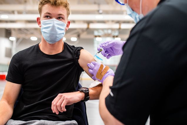 Photo: Mackenzie Fallon of Student Health Services gives a vaccine to Alexander Bade (MET 23) during an immunization clinic on January 23, 2023. A young white person wearing a light blue face mask and black shirt sits in a chair and holds their arm out as a healthcare professional wearing a black collared shirt and light purple surgical gloves gives them a shot.