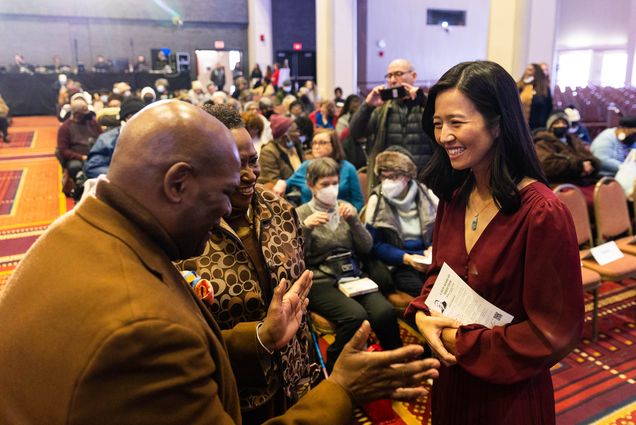 Photo: Boston Mayor Michelle Wu, right, met briefly with former BU Dean of Students Kenneth Elmore, left, and former Howard Thurman Center Director Katherine Kennedy shortly before Boston University, the city of Boston, and the New England Conservatory of Music hosted a joint observance of MLK Day honoring Martin Luther King, Jr. An Asian woman in a burgundy suit ensemble smiles as she looks on to a bald Black man wearing a tweed suit jacket talking and laughing and a shorter Black woman wearing a brown print jacket, also smiling. A blurry crowd of people can be seen seated behind them.