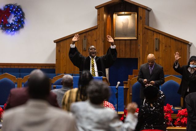 Photo: Reverend Arthur Gordon delivers his sermon at St. John Missionary Baptist Church in Roxbury. A black man wearing a black robe over a white collared shirt and yellow striped tie raises both hands as he gives a sermon at the front of a church.