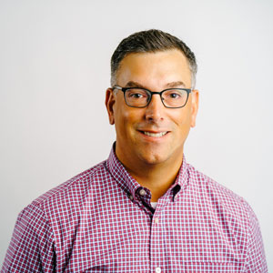 Shea Cronin, a white man wearing glasses and a collared shirt smiles for the camera in front of a white background