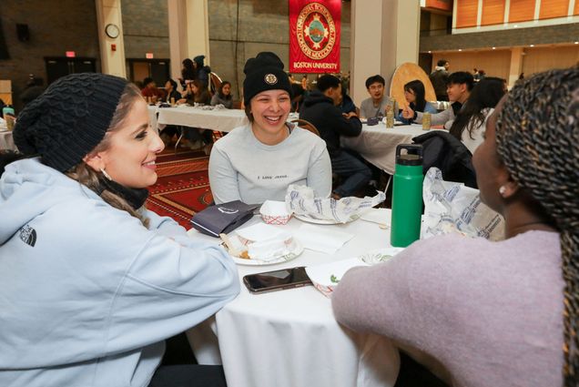 Photo: Maria Zabaneh (CAS’23) (left), Josette Wynn (ENG’25) (middle), and Deirdre Otoo (CAS’23) (right) enjoy their meals together at the Boston Food Fest Jan 21. Three young women sit around a table with a white table cloth as they smile and laugh. Empty white plates sit in front of them on the table and other students can be seen in the background at other, similar tables.