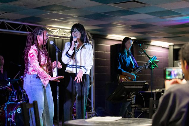 Photo: Sydney Villegas (CAS’24), left, and Stella Lee (CFA’24), right, take the stage together at Karaoke Night on January 28. A young Black woman with long box braids and wearing a pink blouse and jeans, and a young Asian woman wearing a white sweater and black pants, sing into microphones on a stage as a band can be seen playing guitar and drums in the background.