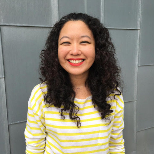 Anna Lim, a young woman wearing a yellow striped shirt smiles happily in this photo in front of a grey background