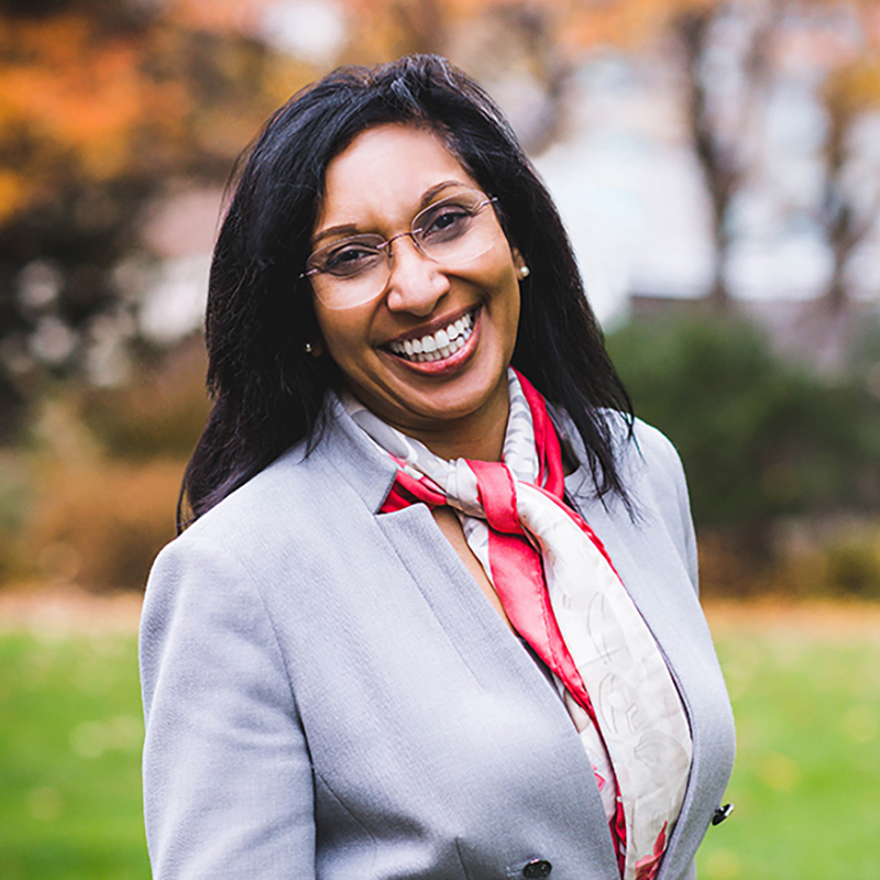 Photo: Headshot of Amanda Bailey, a Black woman with long straight black hair, wearing a white blouse, light blue gray blazer, and red neck scarf, smiles and poses for the camera.