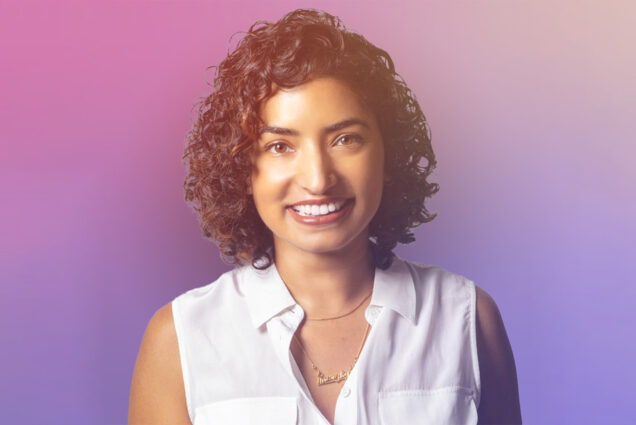 Photo: A watercolor-esque background with pink, blue, and purple. In the front, a feminine-presenting individual with short, curly, brown hair. She is wearing a collared white tank top with two, thin gold chains. She also has s nose piercing and she is smiling in the photo.