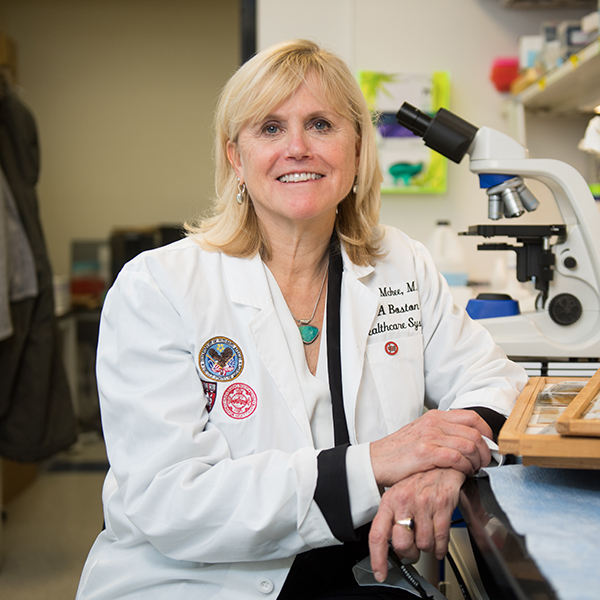 Photo: Dr. Ann McKee, a white woman with shoulder-length blonde hair wearing a white lab coat, sits and poses in a lab next to a large microscope.