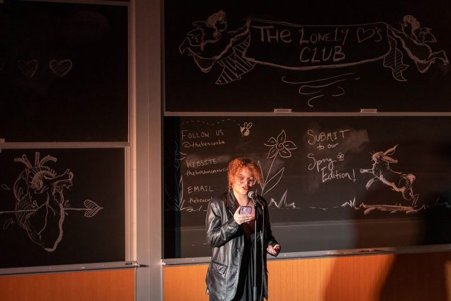 Photo: A young student with curly orange hair and wearing a large leather jacket and black dress reads from her phone as she speaks into a mic set up in front of a blackboard. Behind her, written in chalk on the large blackboard is "The Lonely Club" as well as contact information for the club.