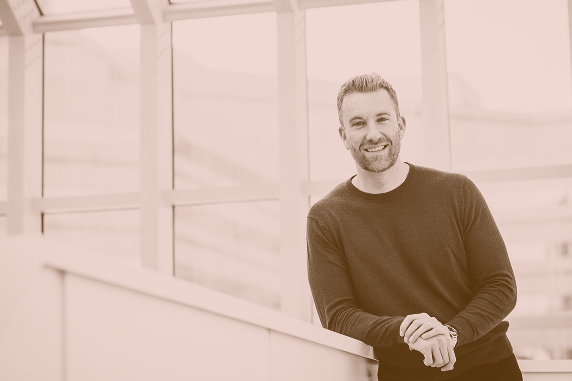 Photo: Arthur Emma, a white man, stands in the right-most part of the frame with a color filter over the photo. His hands are over top one another and he poses, smiling. He is leaning on a railing.