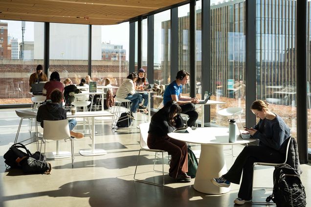 Photo: A sunny shot of the BU Faculty of Computing & Data Sciences Building as students sit and study at modern, round, white tables.